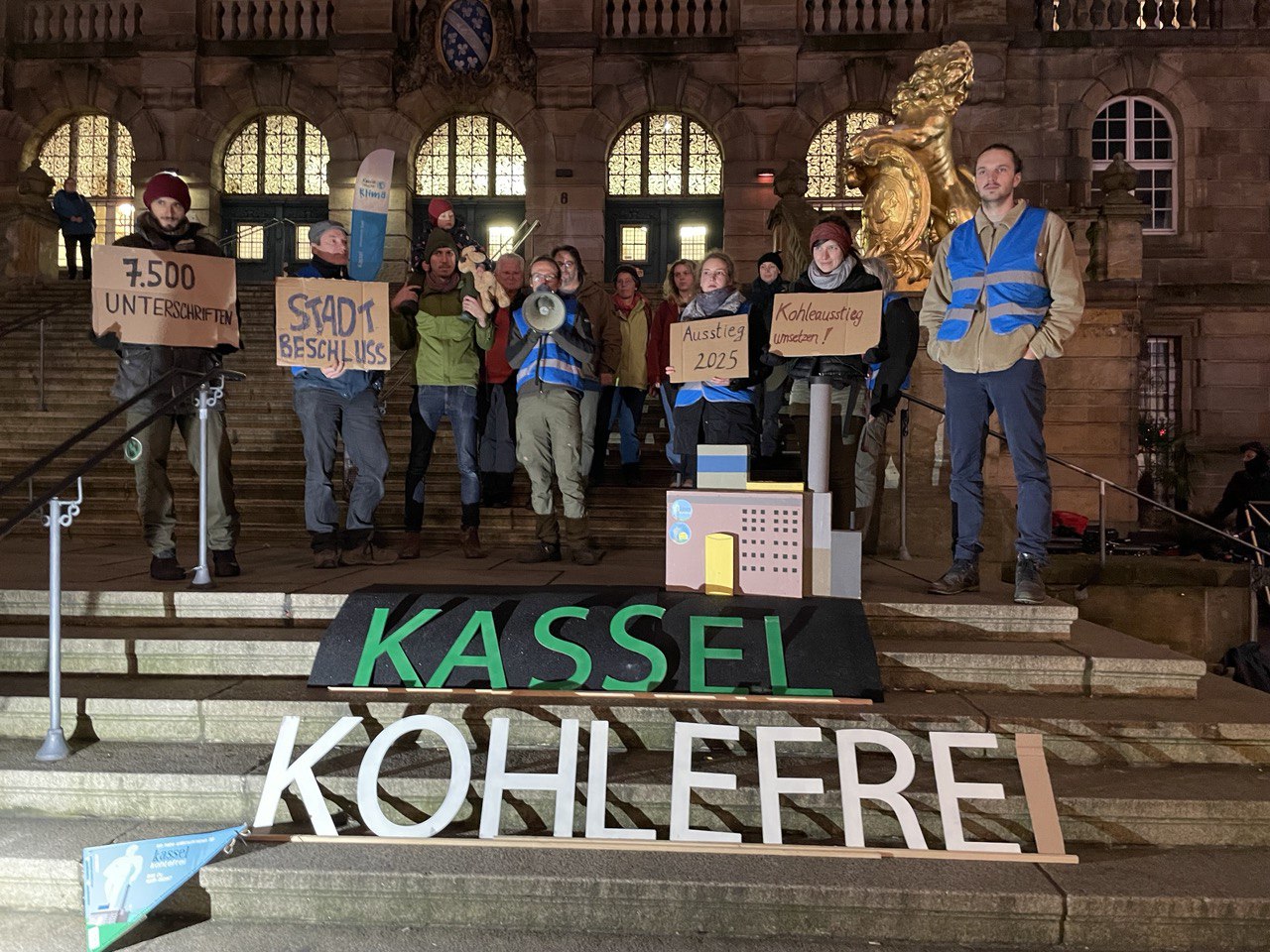 Aktive des Bündnisses kassel kohlefrei mit Westen vor dem Schriftzug auf dem Boden "KASSEL KOHLEFREI" und einem stylisierten Kraftwerk. Im Hintergrund das Kasseler Rathaus mit seinen goldenen Löwen.